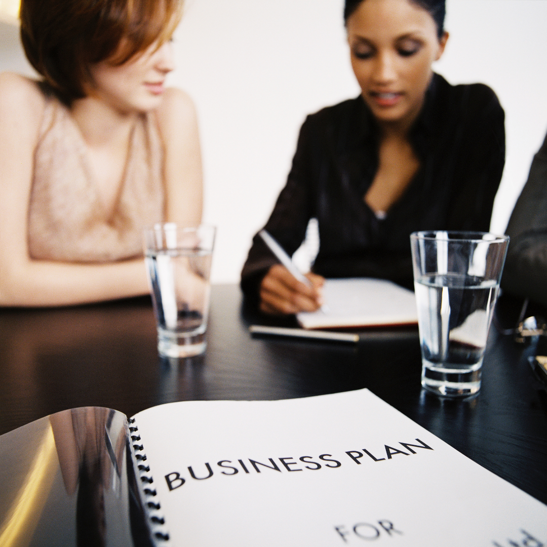 two women sat at desk with two glasses of water discussing business plan document
